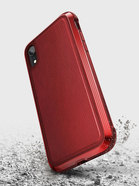 The red iPhone XR Case - LUX by Raptic features a textured back, crafted from impact-resistant polycarbonate. It stands elegantly on a rough grey surface, with small dust particles shimmering around it.