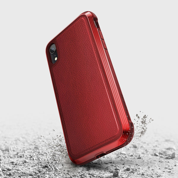 The red iPhone XR Case - LUX by Raptic features a textured back, crafted from impact-resistant polycarbonate. It stands elegantly on a rough grey surface, with small dust particles shimmering around it.