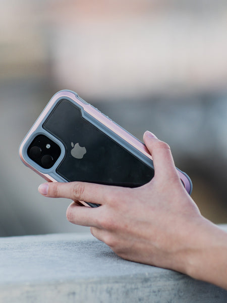A hand holding an iPhone 11 Pro with a dual-camera setup and an Apple logo, encased in a Raptic SHIELD clear protective case made from aluminum alloy, outdoors.