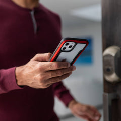 A man holding an iPhone 12 Pro Max with a Raptic SHIELD case in front of a door, ensuring foot drop protection.