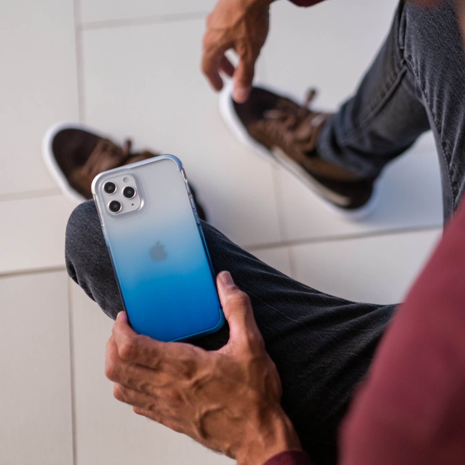 A man is sitting on the floor holding a blue Raptic iPhone 12 Mini Case - Air with drop protection.