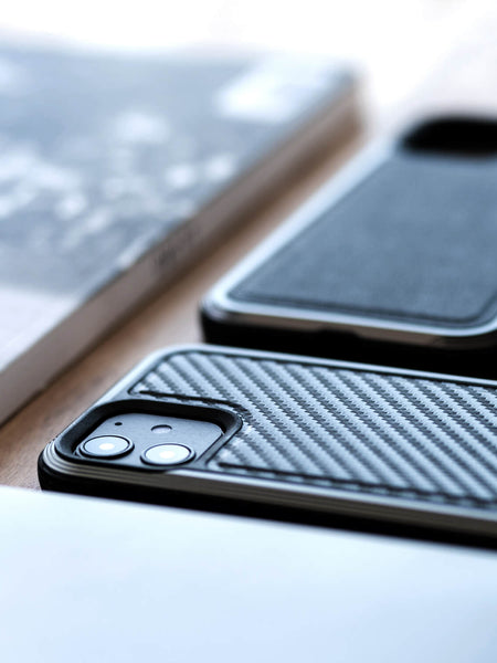 Close-up of two smartphones with black cases, including an iPhone 12 Pro Max adorned in a Raptic iPhone 12 Pro Max Case - LUX for superior drop protection, one showing dual camera lenses, placed on a light-colored surface beside a blurred book or magazine.
