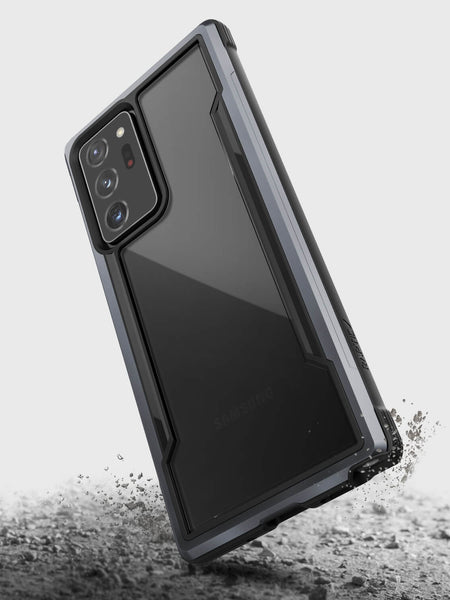 A Raptic Galaxy Note 20 Ultra Raptic Shield case in black is shown protecting a Samsung smartphone as it falls onto a dusty surface, scattering debris—the military-grade drop protection keeps your device safe while still allowing for wireless charging.