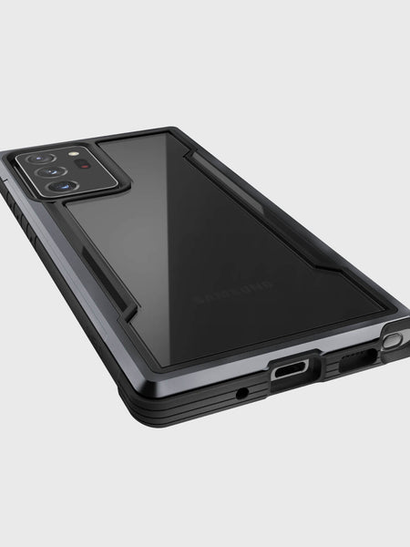 A Galaxy Note 20 Ultra fitted with a black Raptic Shield case, featuring military-grade drop protection, is displayed lying face down on a plain, light gray surface.