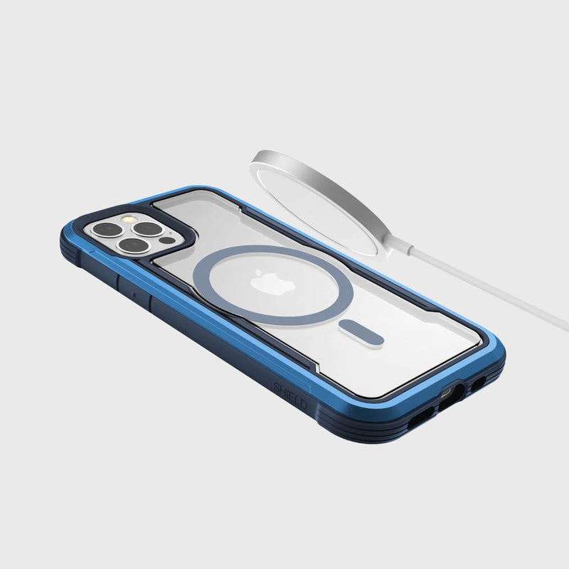 The Raptic SHIELD PRO MAGNET case for iPhone 12 & 12 Pro snugly fits, showcasing the rear camera and Apple logo. Nearby, a MagSafe charger promises effortless charging on a light grey background while providing reliable drop protection for your device.