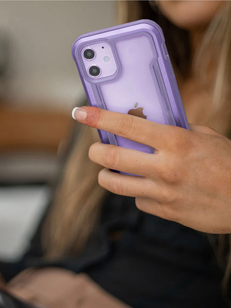 Indoors, a person holds an iPhone 12 with a Raptic Shield case in purple. The sleek machined aluminium bumper offers military-grade drop protection and wireless charging compatibility, blending style with peace of mind.