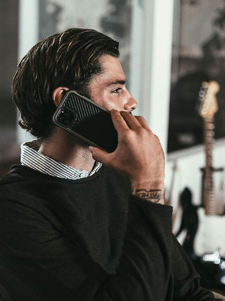 A man with a wrist tattoo holds a smartphone to his ear, the Raptic LUX iPhone 11 Pro Max Case glinting in the light, as he sits indoors near a guitar.