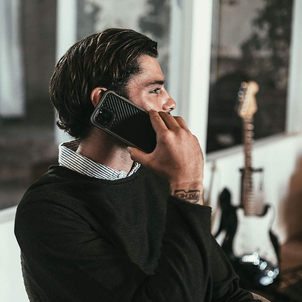 A man with a wrist tattoo holds a smartphone to his ear, the Raptic LUX iPhone 11 Pro Max Case glinting in the light, as he sits indoors near a guitar.