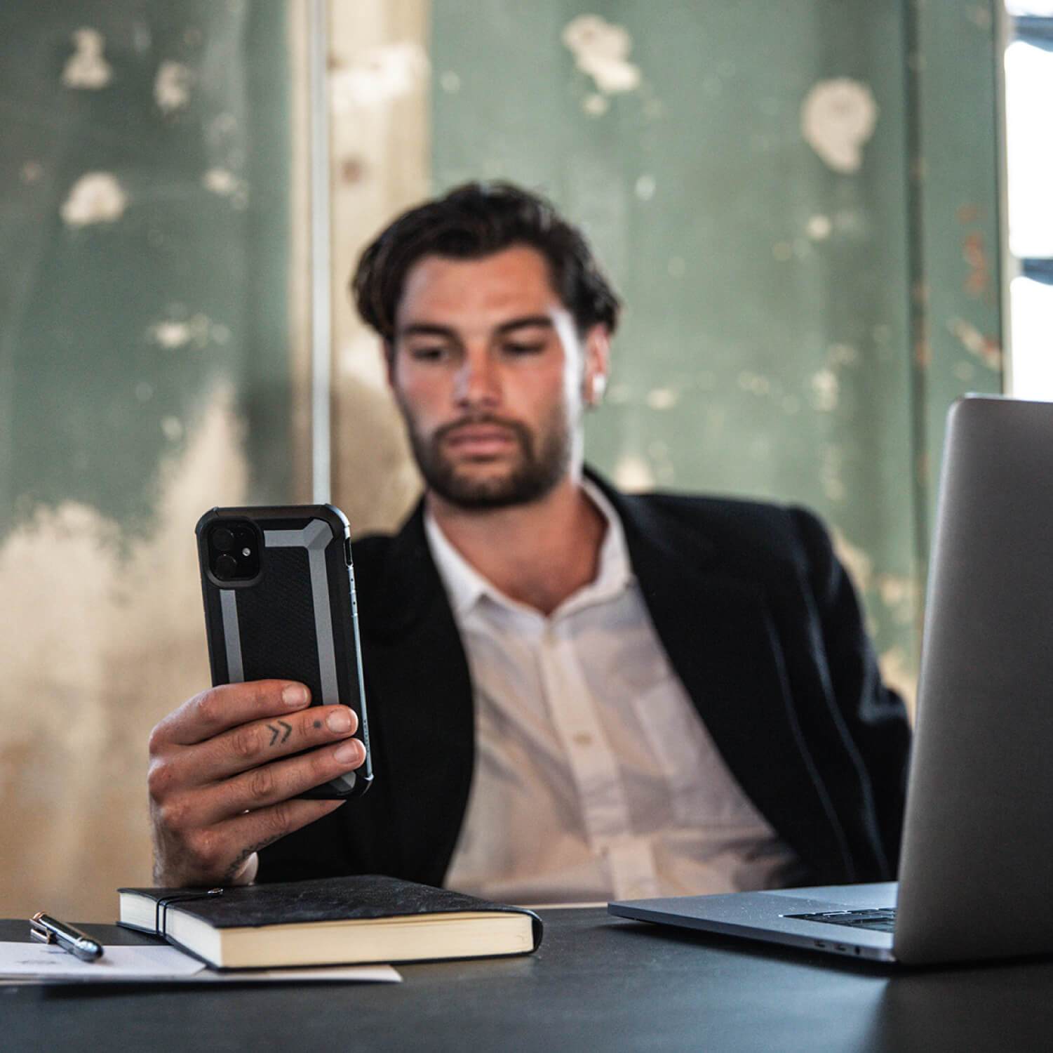 A man sitting at a desk with a laptop and a cell phone, protected by a shock-absorbing rubber exterior Raptic iPhone 11 Pro Max Case - TACTICAL that meets Military Standard MIL-STD-810.