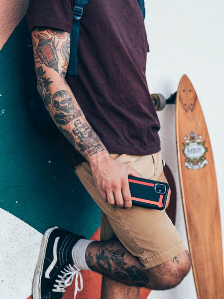 A man with tattoos leaning against a wall holding a skateboard while showcasing his Raptic Tactical protective iPhone 11 Pro Max case. This durable case features a shock-absorbing rubber exterior, meeting the TACTICAL brand's high standards for quality and protection.