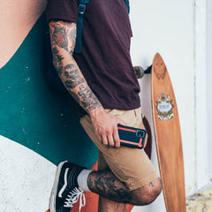 A man with tattoos leaning against a wall holding a skateboard while showcasing his Raptic Tactical protective iPhone 11 Pro Max case. This durable case features a shock-absorbing rubber exterior, meeting the TACTICAL brand's high standards for quality and protection.