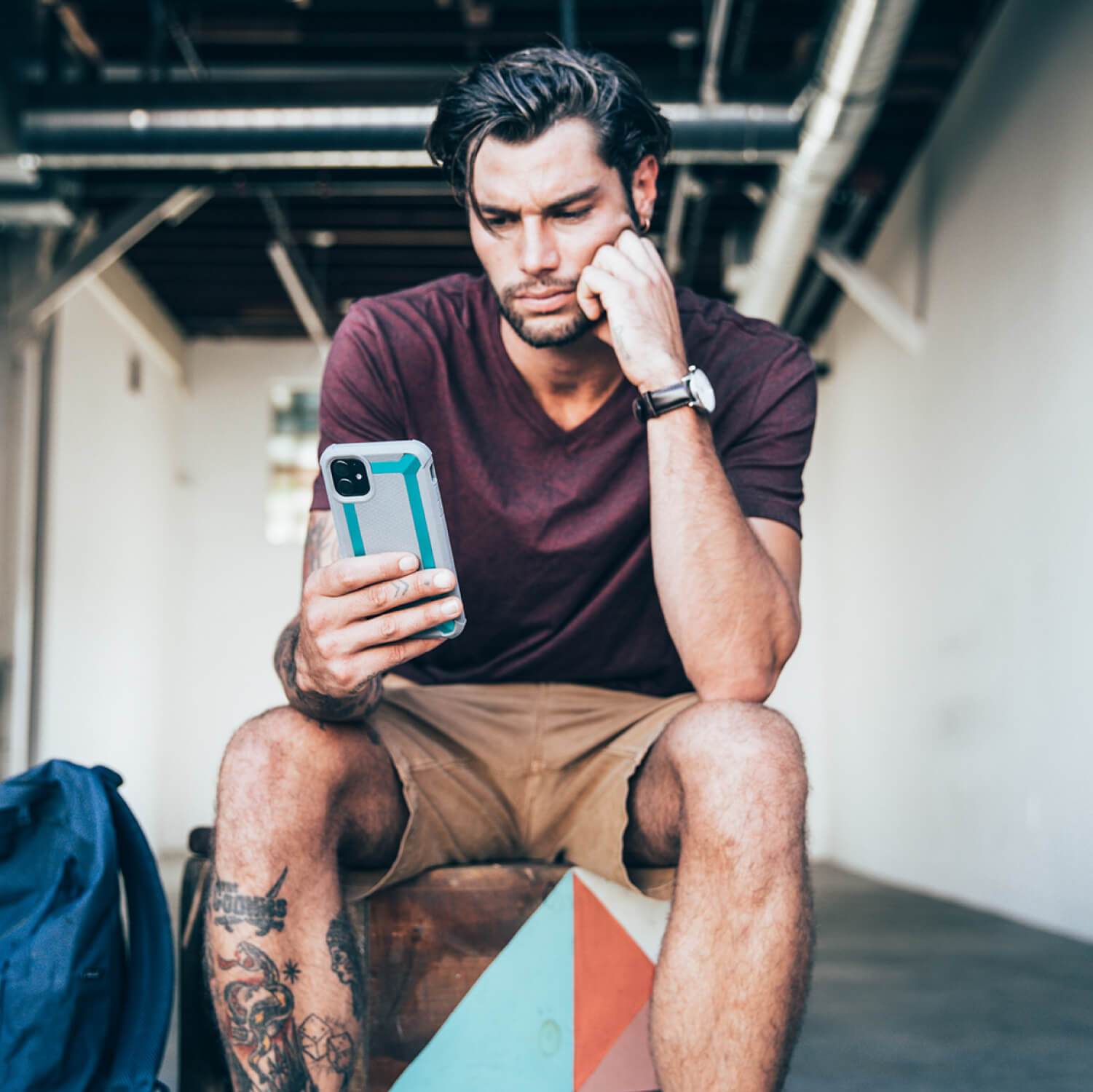 A man sitting on top of a box, browsing his phone while using a Raptic shock-absorbing protective case for his iPhone 11 Pro.