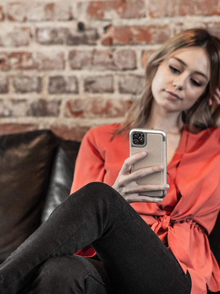 A woman is sitting on a couch looking at her iPhone 11 protected by a Raptic PRIME case.