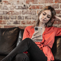 A woman is sitting on a couch looking at her iPhone 11 protected by a Raptic PRIME case.