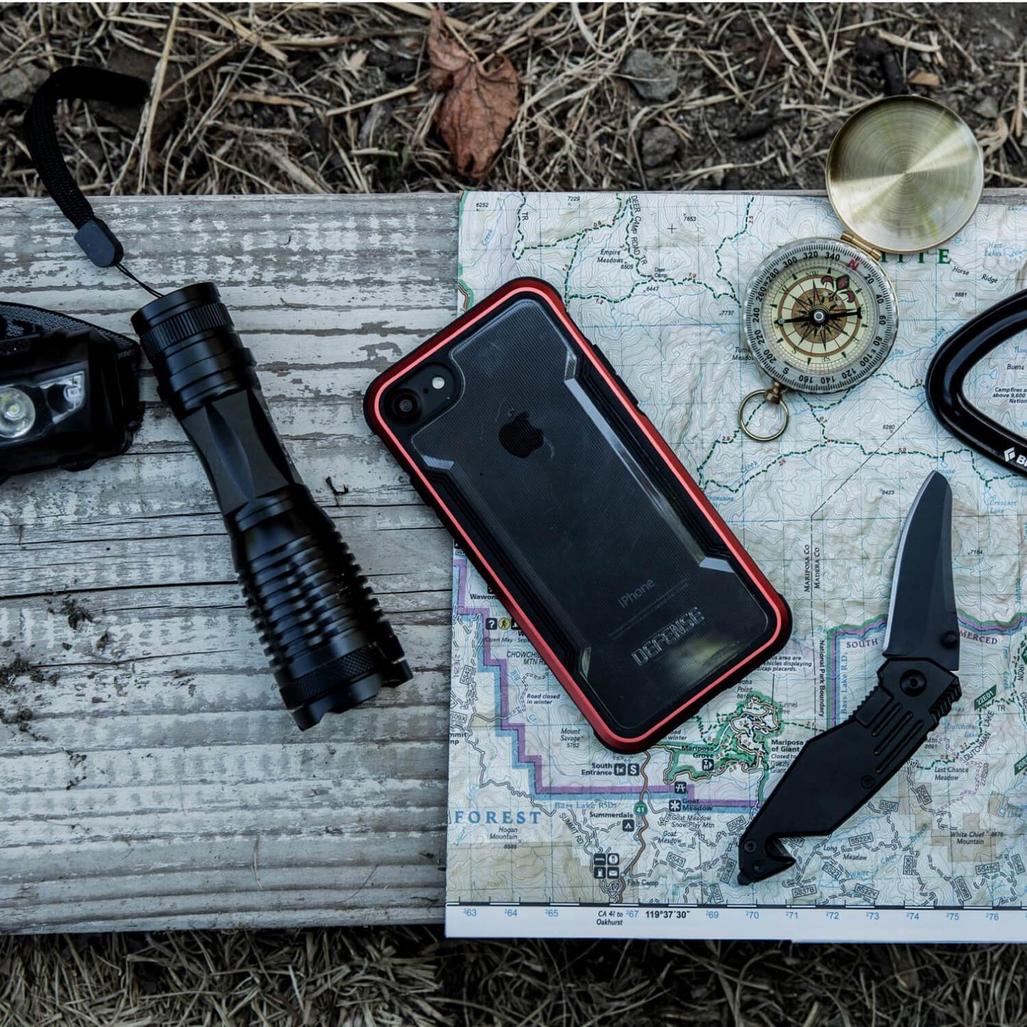 A map, a knife, a flashlight, and an iPhone SE/8/7 Case - SHIELD by Raptic are on a wooden table.