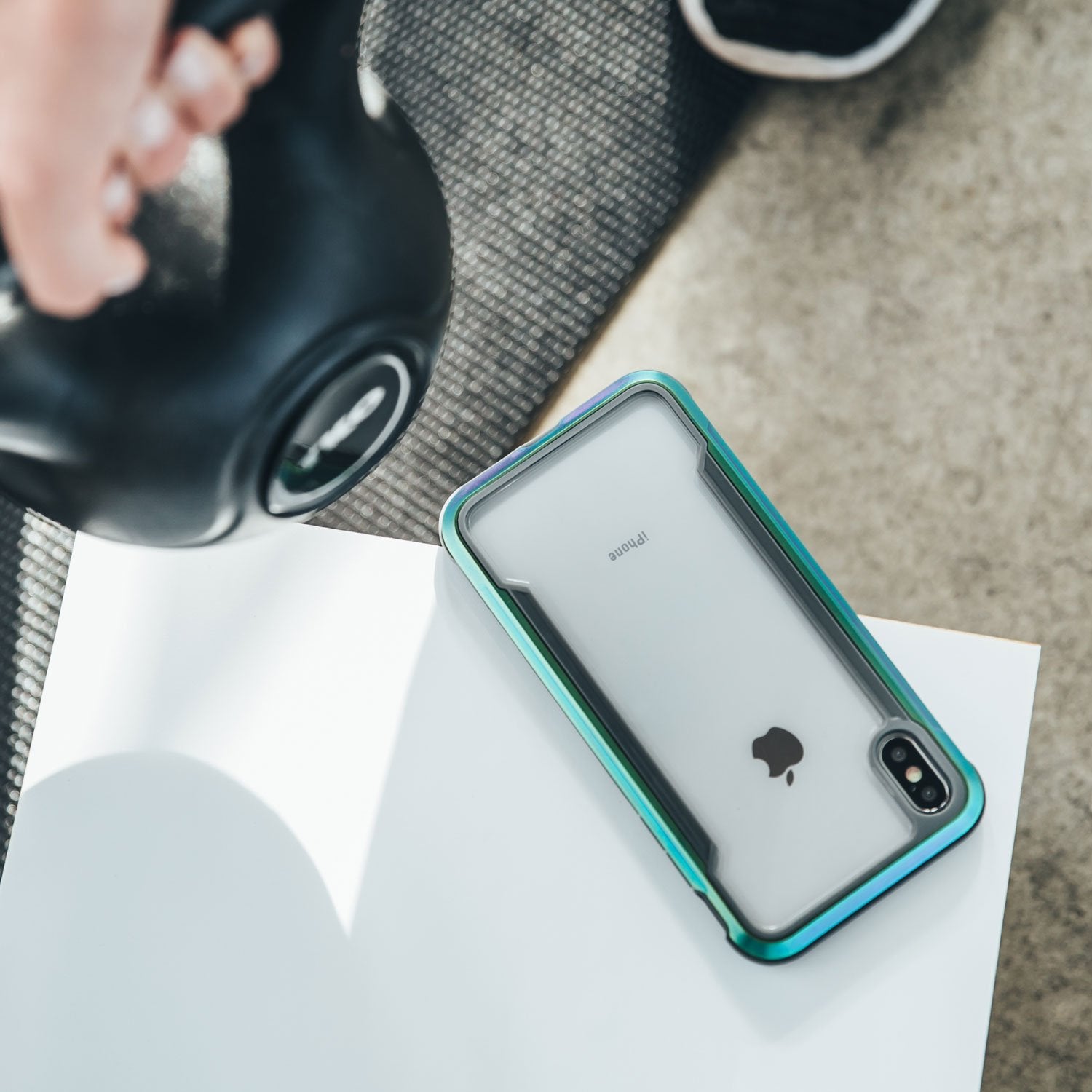 A person is holding a Raptic SHIELD iPhone XS Max Case-protected iPhone X/XS on a table next to a kettlebell.