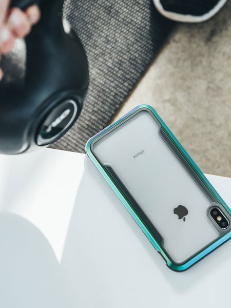 An iPhone X proudly wrapped in a Raptic SHIELD case rests on a white surface, offering military-grade drop protection, while someone nearby confidently holds a black kettlebell.
