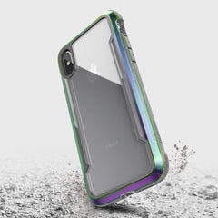 A smartphone with a Raptic iPhone X/XS Shield case in metallic green and purple floats above a textured surface, highlighting its military-grade drop protection.