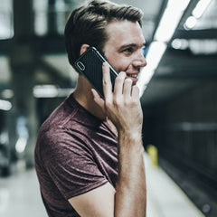 In a bustling subway station, a person in a maroon shirt cheerfully talks on their iPhone XS Max, protected by the impact-resistant Raptic LUX case.