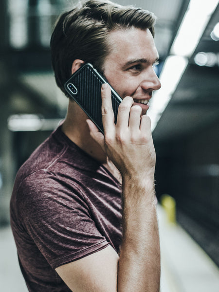 In a bustling subway station, a person in a maroon shirt cheerfully talks on their iPhone XS Max, protected by the impact-resistant Raptic LUX case.