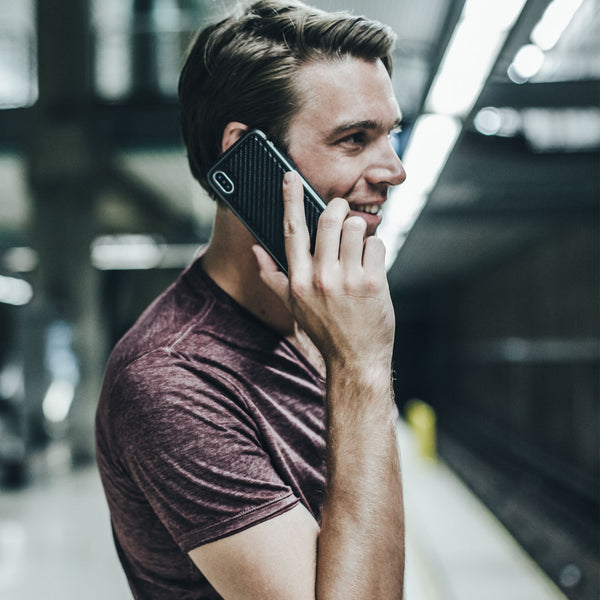In a bustling subway station, a person in a maroon shirt cheerfully talks on their iPhone XS Max, protected by the impact-resistant Raptic LUX case.