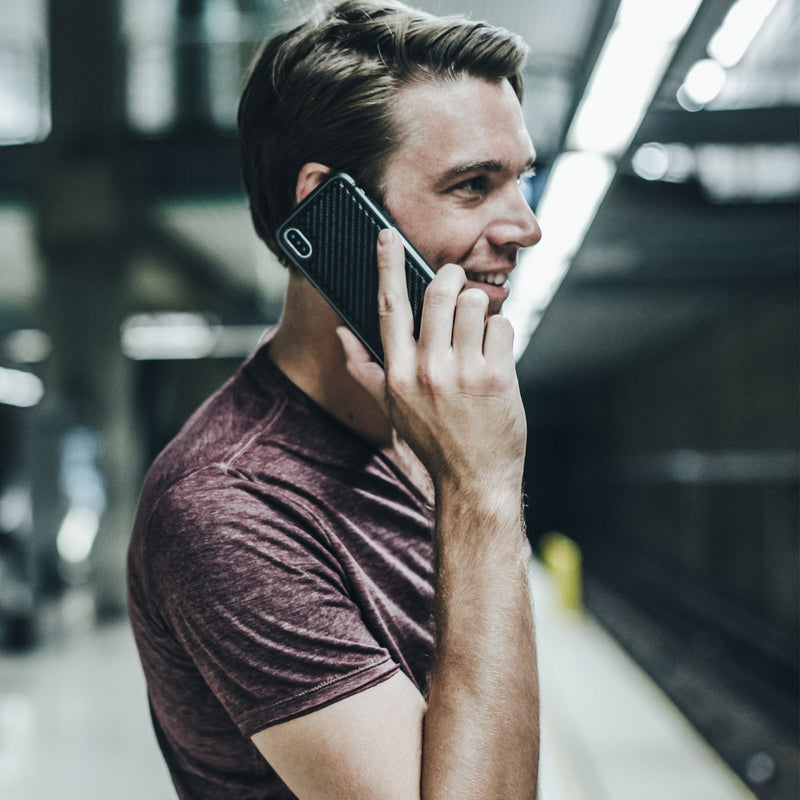 In a bustling subway station, a person in a maroon shirt cheerfully talks on their iPhone XS Max, protected by the impact-resistant Raptic LUX case.